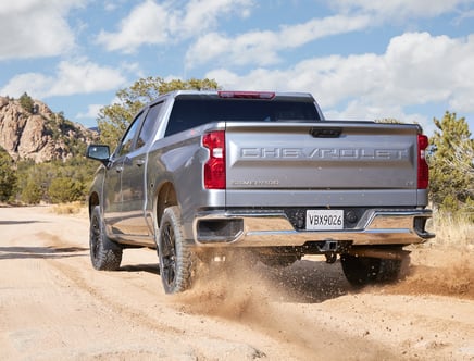 A silver Chevrolet Silverado driving on a dirt road in a desert landscape.