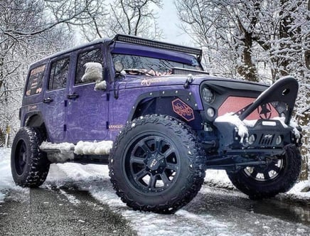 A purple Jeep Wrangler with large off-road tires and custom modifications, parked on a snowy road with snow-covered trees in the background.