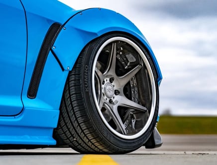 Close-up of a blue car's front wheel with a silver alloy rim and a textured black air vent on the fender.