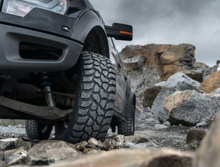 Close-up of a black off-road vehicle navigating over large rocks with rugged terrain and cloudy sky in the background.