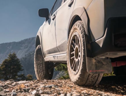 Close-up of a rugged vehicle's tire on a rocky path with mountains in the background.