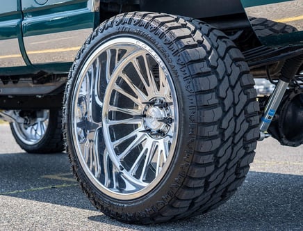 A large truck tire with chrome alloy rim on an asphalt surface.