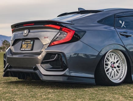 A modified gray Honda Civic Si with custom wheels and rear spoiler parked on grass.