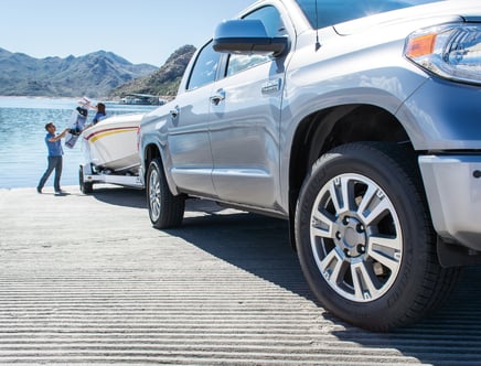 A pickup truck towing a boat on a trailer at a boat ramp near a lake, with mountains in the background.