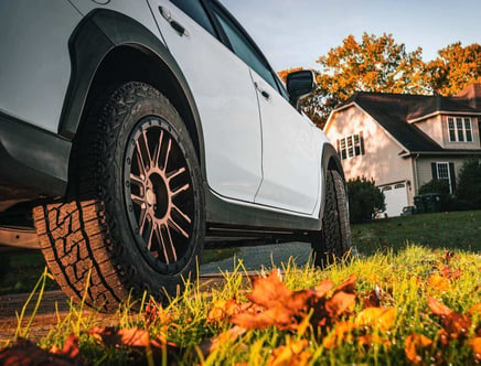 Low angle view of a white SUV with rugged tires parked in front of a house during autumn, with fallen leaves on the grass.