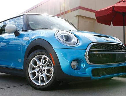 Blue Mini Cooper with reflections of the sky and trees, parked near a building with a red umbrella.