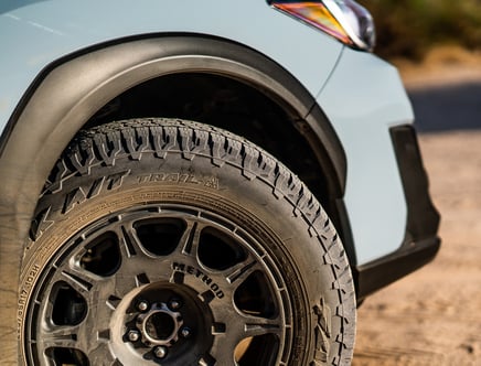 Close-up of a car's front tire with an all-terrain tread pattern and black alloy wheel on a sandy surface.