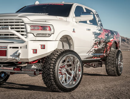 A white lifted pickup truck with large chrome wheels and custom decals on the side, displayed in a desert setting.