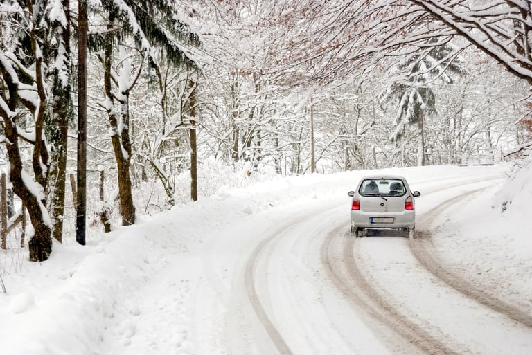 A car driving on a snow-covered road through a forest with snow-laden trees.