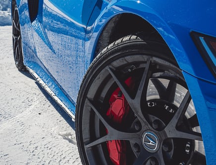 Close-up of a blue sports car's front wheel and fender on a snowy surface, showing a black alloy wheel with a red brake caliper.