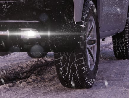 A close-up of a vehicle's tire and wheel on a snowy, icy road, with a bright light shining from the vehicle.