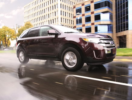 A dark-colored SUV driving on a wet city street with office buildings in the background.