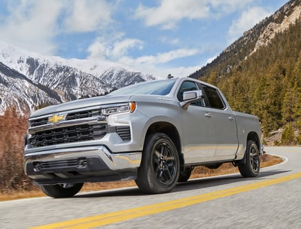 Silver pickup truck driving on a mountain road with snowy peaks and pine trees in the background.