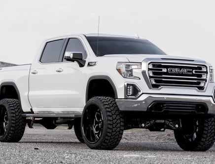 A white lifted GMC truck with large black wheels parked on a gravel surface.