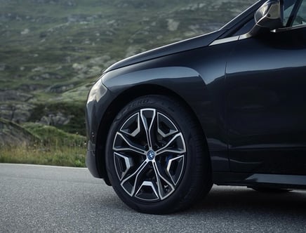 Front wheel and fender of a dark-colored car on a road with a grassy landscape in the background.