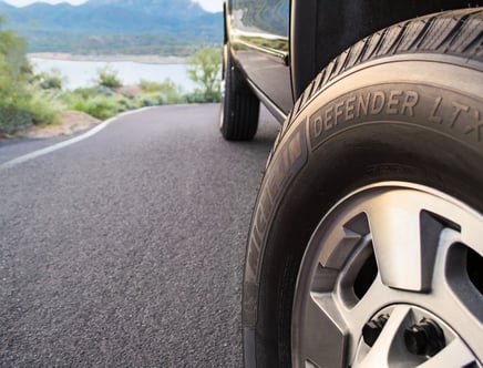 Close-up of a Michelin Defender LTX tire on a vehicle, with a winding road and scenic landscape in the background.