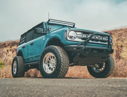 A lifted teal blue off-road SUV with large tires and a front bumper bar is parked on a rural road with dry grass and hills in the background.