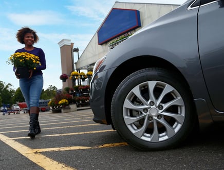 A woman carrying a pot of yellow flowers walks in a parking lot next to a gray car.