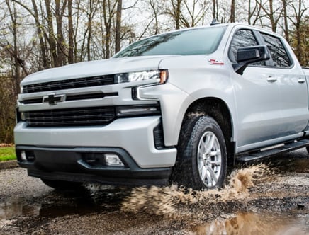 A white pickup truck driving through a muddy puddle on a wet road with trees in the background.