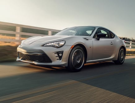 A silver sports car driving on a road with a white fence in the background.