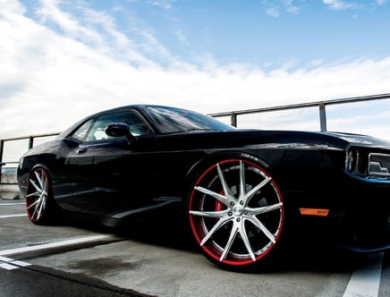 A black sports car with custom red and silver alloy wheels parked in a parking lot under a cloudy sky.