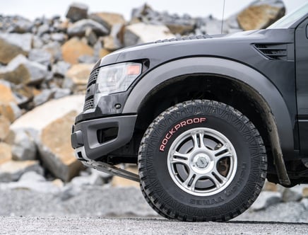 Close-up of a black off-road vehicle's front tire with "ROCKPROOF" marked on it, against a rocky background.