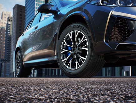 Close-up view of a car's front wheel with black and silver rims, featuring Dynapro HPX tires, set against a city background.