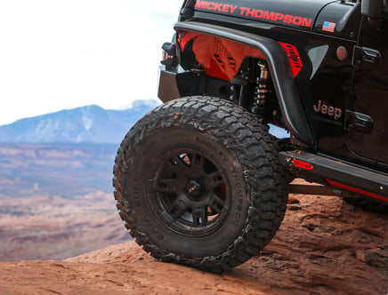 Close-up of a Jeep with a Mickey Thompson tire on rocky terrain, with a mountainous landscape in the background.