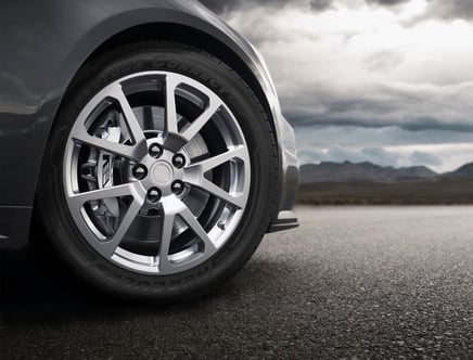 Close-up of a car wheel with alloy rim on a highway, with a cloudy sky and mountain range in the background.