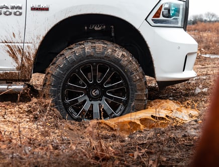 Close-up of a white truck's front wheel and tire partially covered in mud, splashing through a muddy terrain.