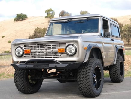 A vintage silver Ford Bronco with large off-road tires, parked outdoors near a grassy hillside.