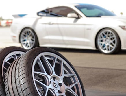 A white sports car parked on a road with a set of alloy wheels and tires in the foreground.