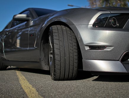 Close-up of a silver sports car parked on the road, emphasizing the front tire and headlight against a clear blue sky.