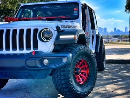 White Jeep Rubicon with red wheels and a red jack mounted on the hood, parked with a city skyline in the background.