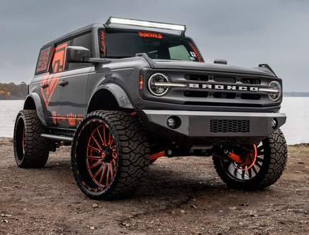 Customized grey Ford Bronco with large wheels and orange accents parked near a body of water.