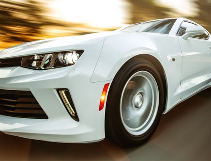 Close-up of a white sports car in motion, highlighting the front wheel and headlight, with a blurred background indicating speed.