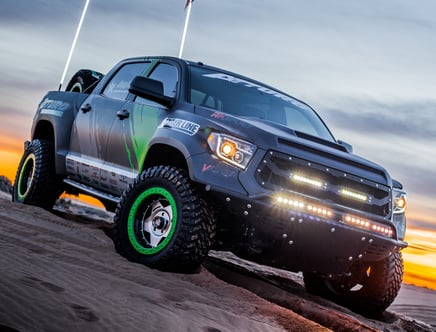 Custom off-road truck with LED light bars and green-accented wheels on a sandy terrain at sunset.