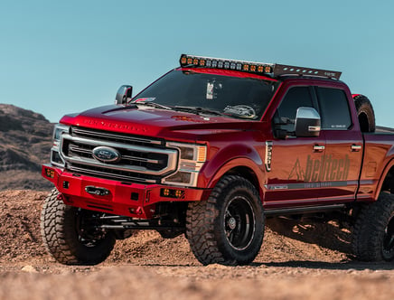 Red Ford Super Duty truck with off-road modifications, featuring large tires and a light bar, parked on rough terrain.