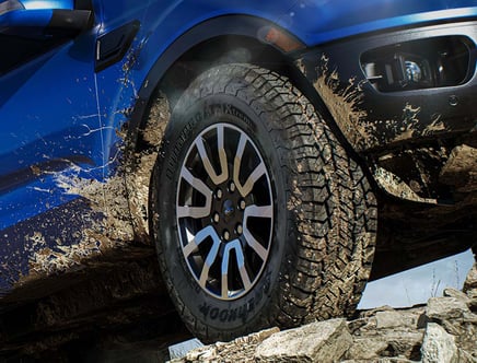 Close-up of a muddy tire on a blue off-road vehicle driving over rocky terrain.