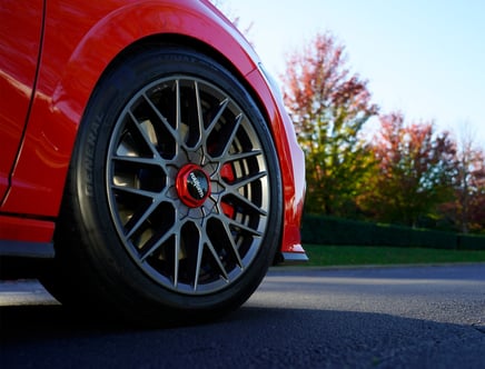 Close-up of a red car's wheel with aftermarket alloy rims on a road, with trees displaying autumn foliage in the background.