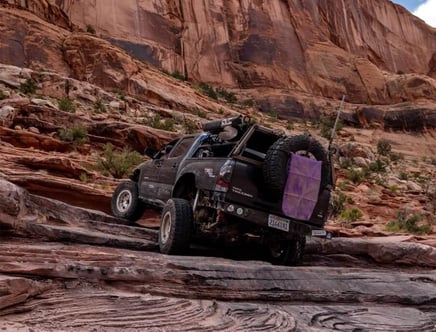 A 4x4 vehicle navigating rocky terrain with steep, layered cliffs in the background.