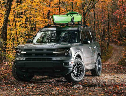 Gray SUV with a kayak on top, parked on a forest trail amidst autumn foliage.