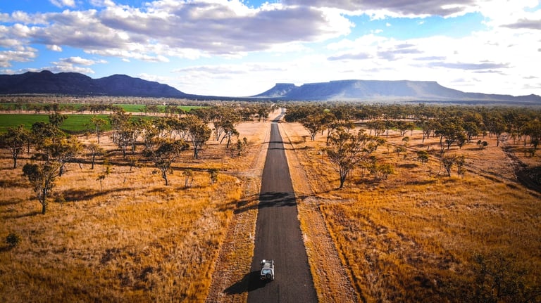 Aerial view of a solitary car driving down a straight road through dry grasslands with distant mountain ranges under a partly cloudy sky.