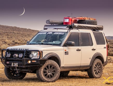 White Land Rover SUV equipped for off-road with roof rack, spare tire, fuel canisters, and camping gear, set against a twilight landscape with a crescent moon.