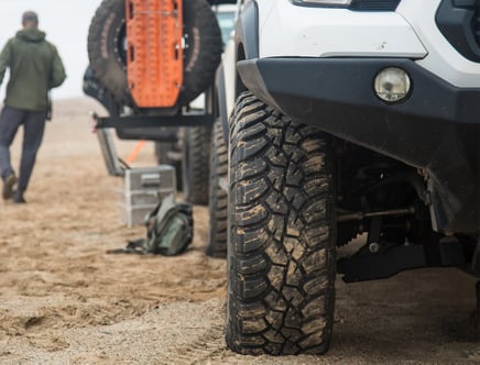 Close-up of an off-road vehicle's tire on a sandy terrain, with recovery gear and a person walking in the background.