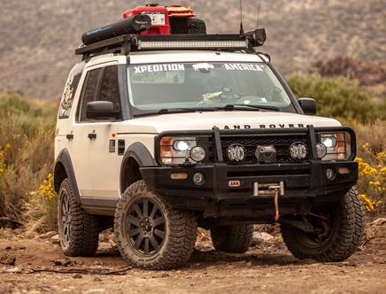 A rugged Land Rover equipped for off-road adventures with roof gear and front-mounted lights, parked on a rocky trail.